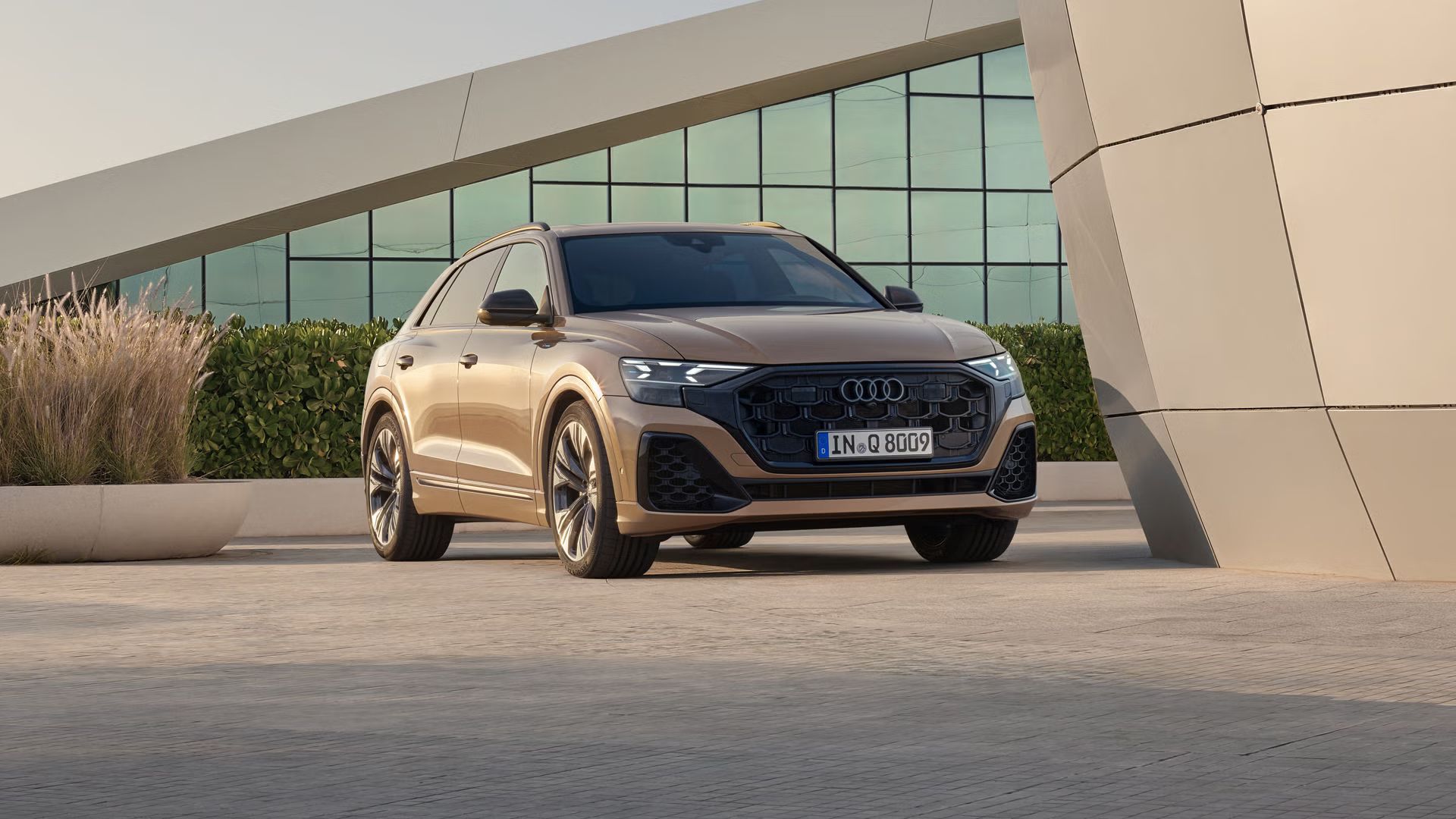 Der Q8 SUV schräg von vorn parkt vor einem modernen Gebäude. Eine bepflanzte Hecke trennt das Fahrzeug von dem dahinter liegenden, voll verglasten Gebäude.
