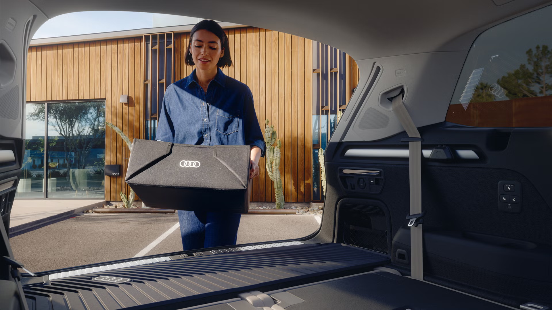 Blick von der Rückbank eines Audi Q7 auf den geöffneten Kofferraum mit Fokus auf die Gepäckraumschale. Eine junge Frau stellt darauf eine kleine Kiste mit Audi-Logo ab.